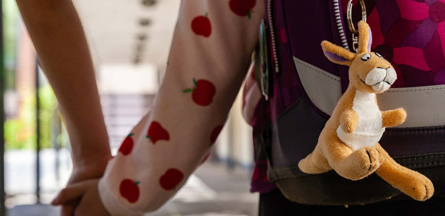 Ein Plüschkänguru hängt am Schulranzen eines Kindes. Das Kind hält Hand mit einem Erwachsenen