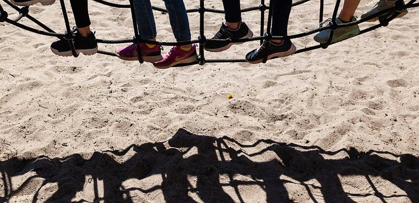 Kinder laufen auf einem Sandspielplatz über eine Hängebrücke. Auf dem Sand sind die Schatten der Kinder zu sehen