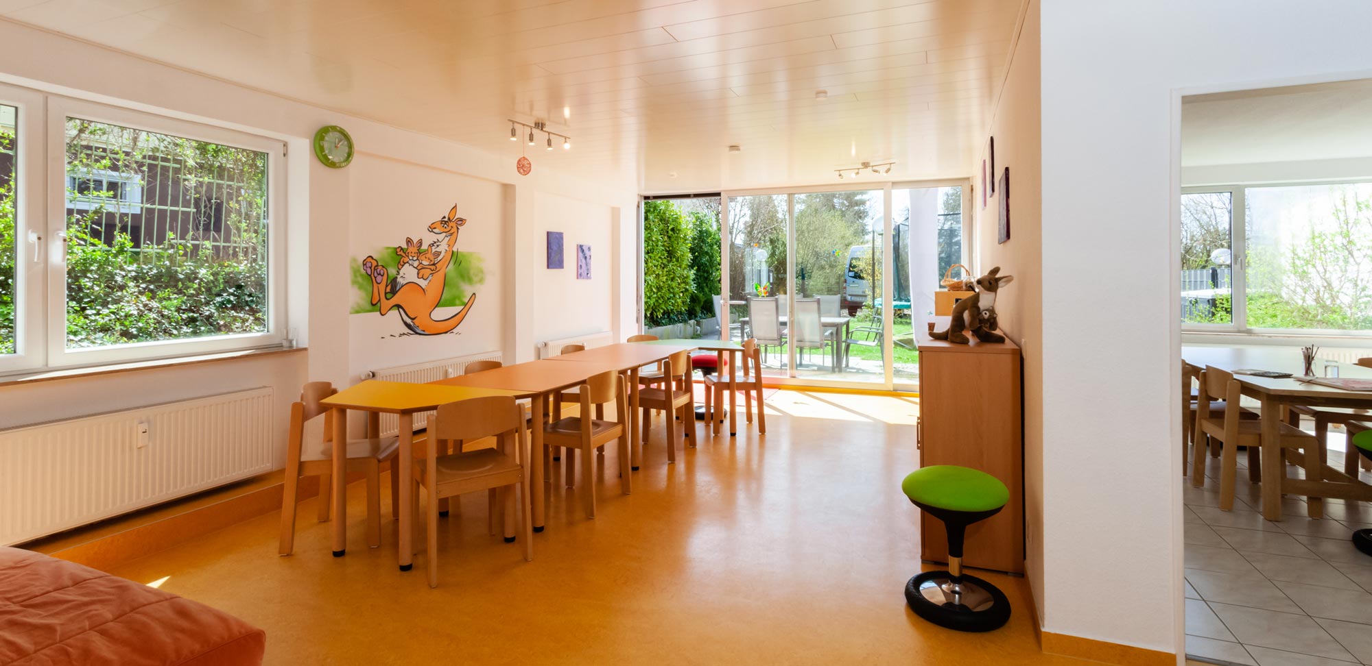 Raumbild. Links ein großer Raum mit einer Tafel und vielen Fenstern. Rechts angeschnitten ein Blick in die Küche. 